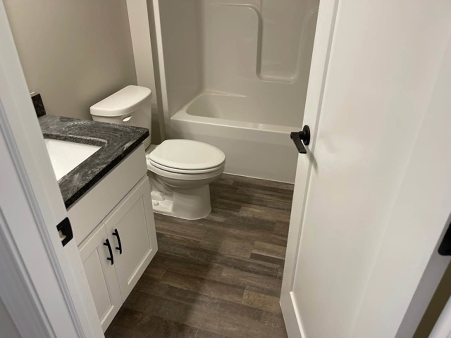 White toilet with closed seat next to a built-in bathtub, wood flooring, light-colored tile walls, and cabinetry in a residential bathroom.