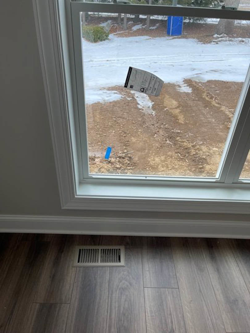 Double-hung window with white trim, a sheet of paper wedged between glass panes, wood floor below, wall vent visible nearby