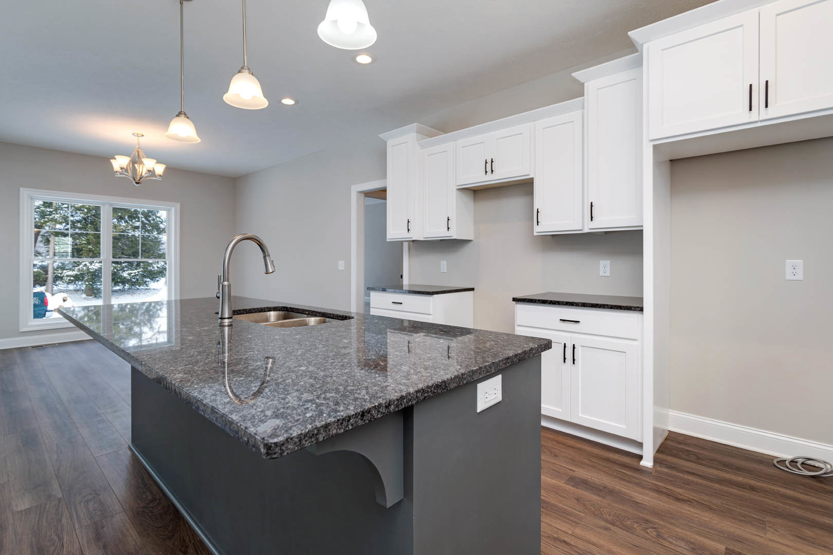 Granite island with undermount sink, white shaker cabinets, stainless steel faucet, pendant chandelier, window overlooking snowy yard, white electrical outlet on backsplash