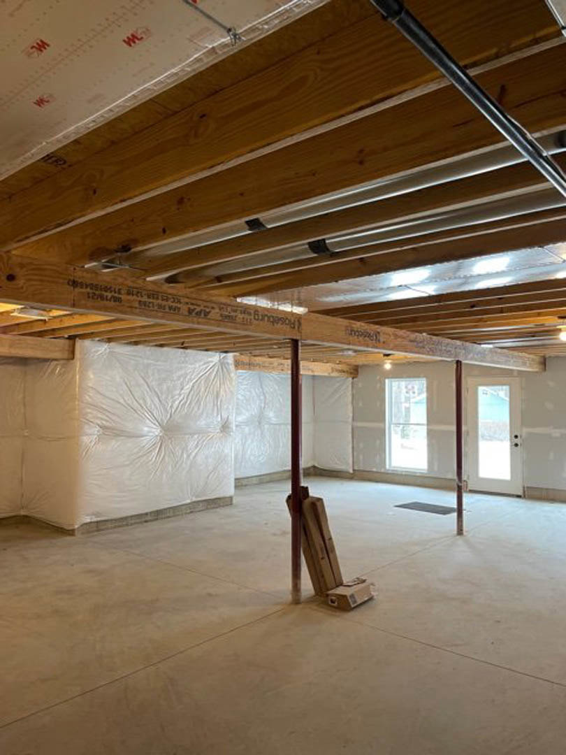Wood-beamed ceiling with exposed pipes, white-framed window, stacked firewood on a stand, cardboard boxes near a support pole, plaster walls, and a wooden door.