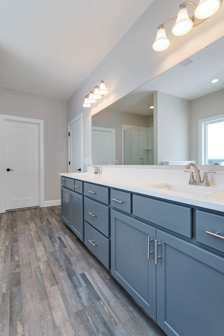 Bathroom with blue shaker cabinets, wide quartz countertop, large frameless mirror, row of wall-mounted lights, white door with black handle, tile flooring, and window with white
