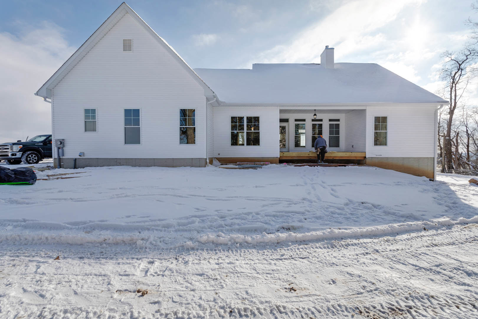 Two-story house with white-framed windows, snow-covered yard with tire tracks, leafless trees, and a person in a blue sweater standing near a parked truck.