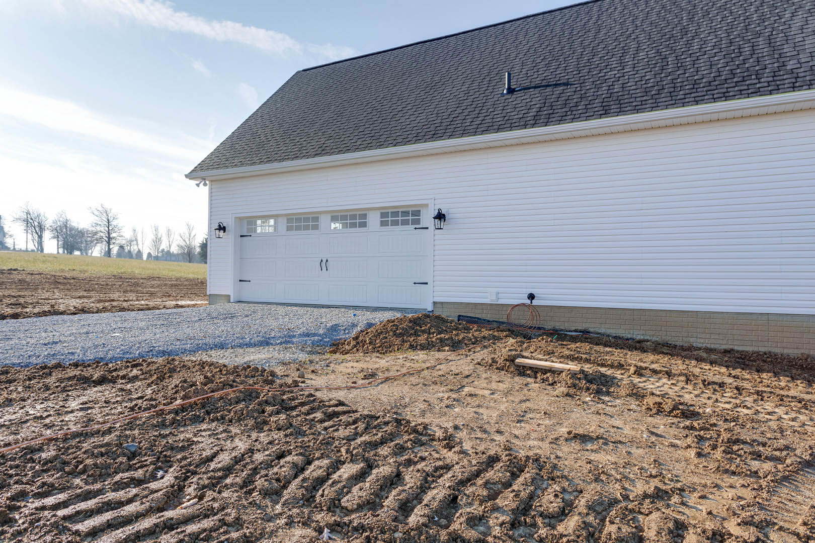 White garage with black roof, dirt patch and piles of soil in front, surrounded by grassy land and trees under cloudy sky