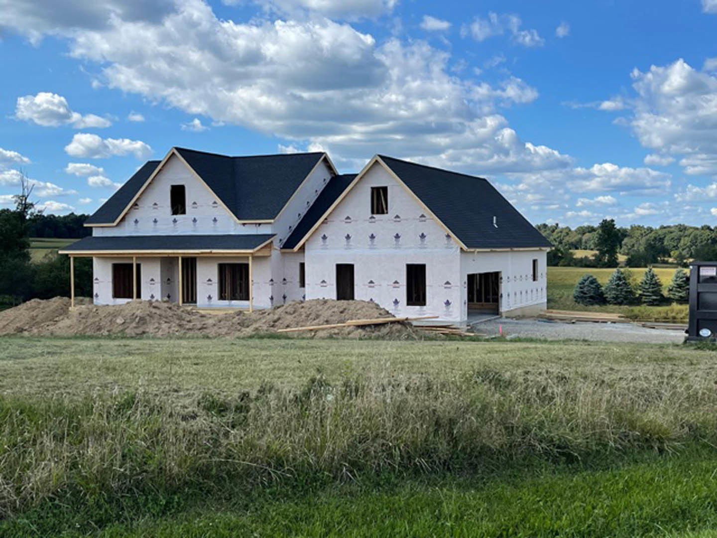 Partially built farmhouse with exposed framing, piles of dirt and wood, grassy field, blue sky with scattered clouds