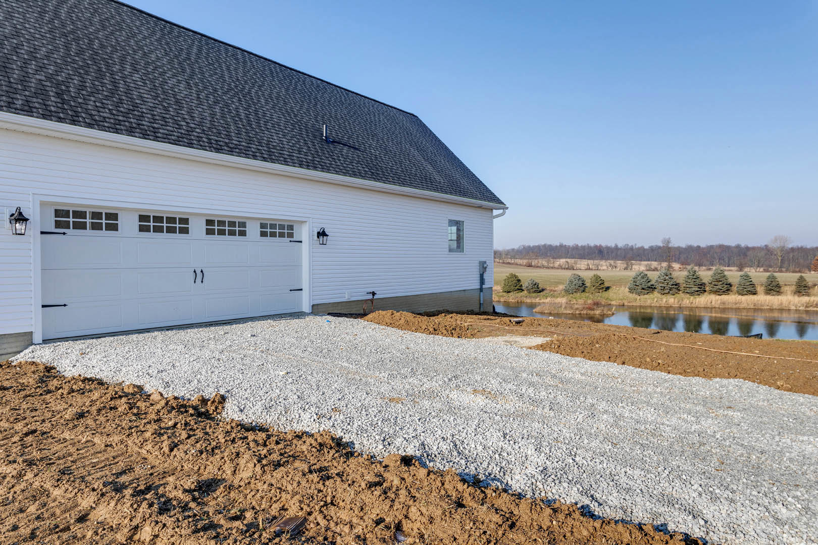 White house with black roof and attached garage, gravel driveway bordered by grass, several trees scattered across open field under blue sky.