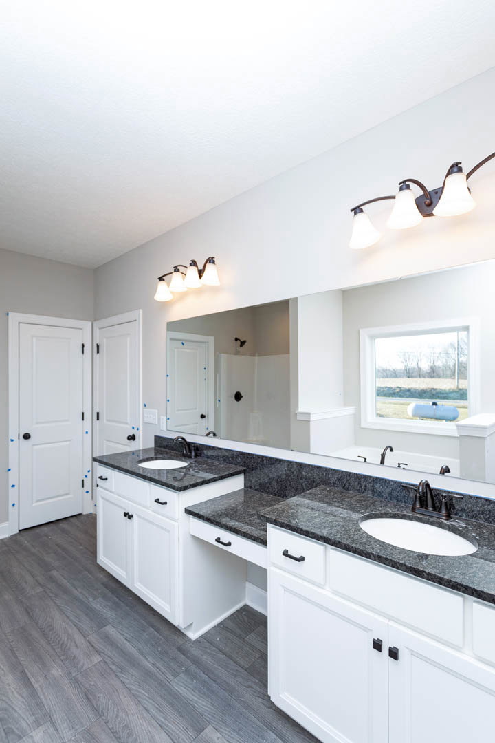 Bathroom with double white sinks set in black granite countertops, large frameless mirror above, white cabinetry with black hardware, window overlooking grassy field, modern light