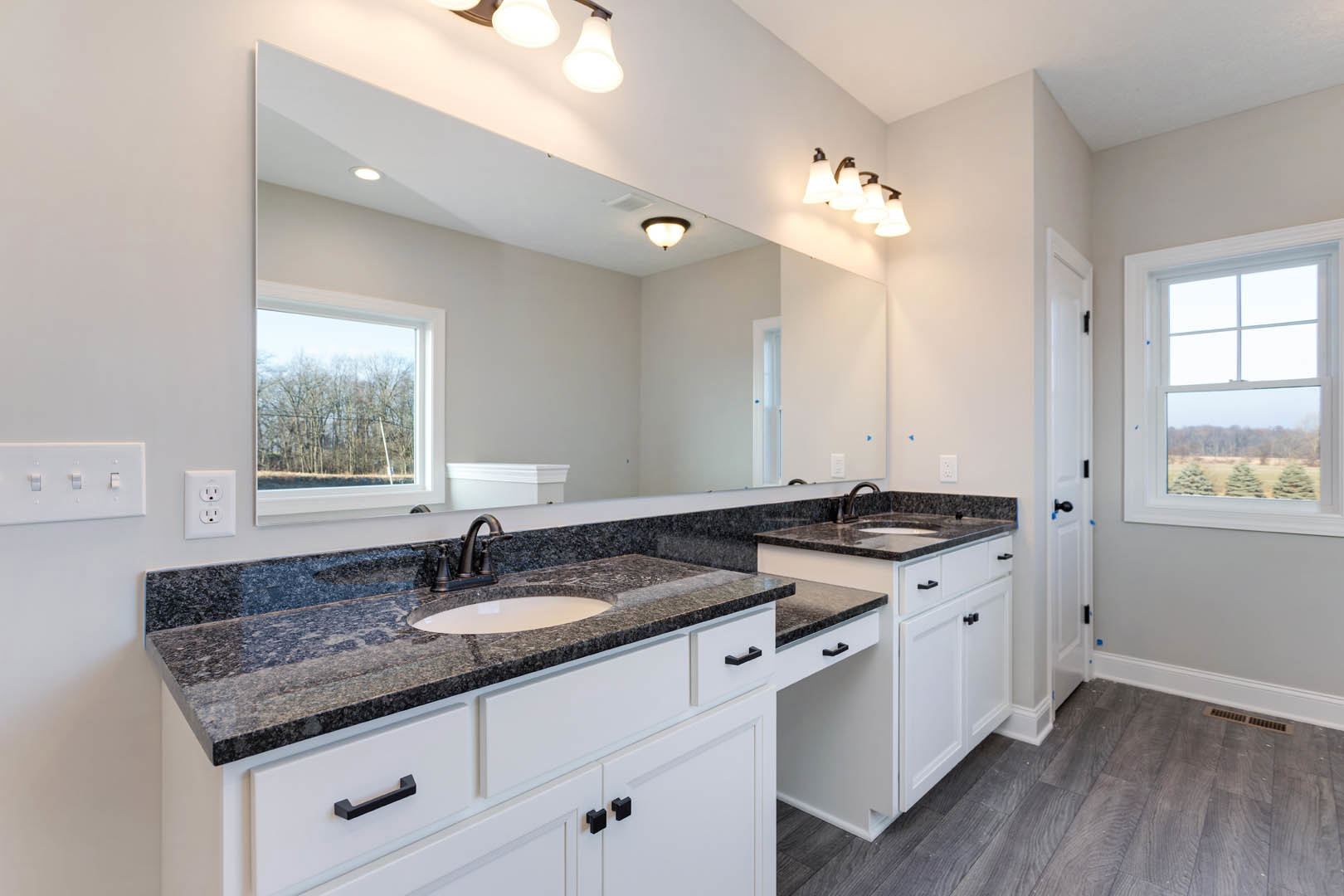 Bathroom with expansive mirror above a tile countertop, modern sink and tap, cabinetry below, window overlooking trees, light fixture mounted on wall, visible outlet near sink.