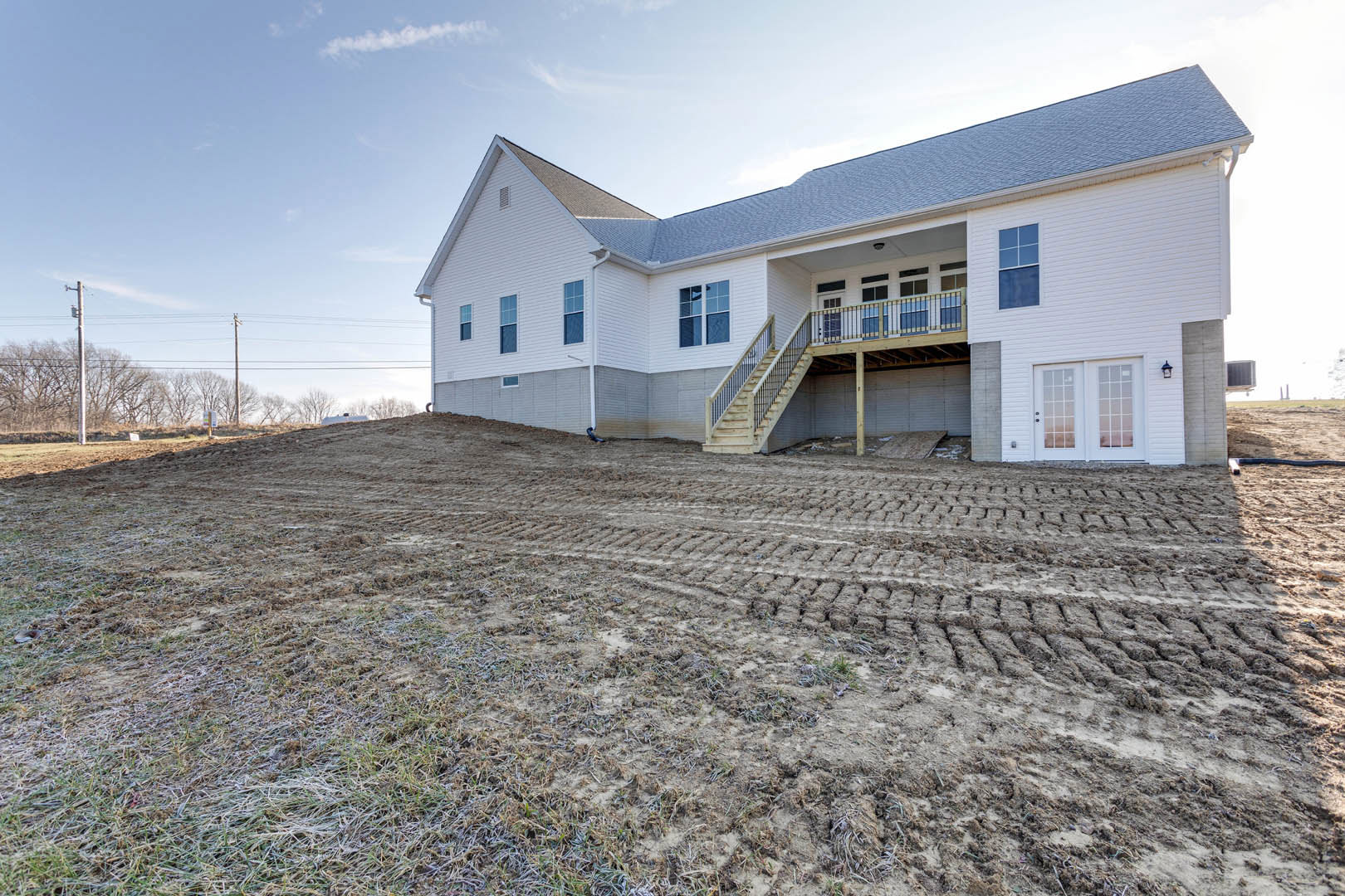 Two-story house with double front doors, large windows, and a staircase leading to the entrance, surrounded by a dirt field under a partly cloudy sky