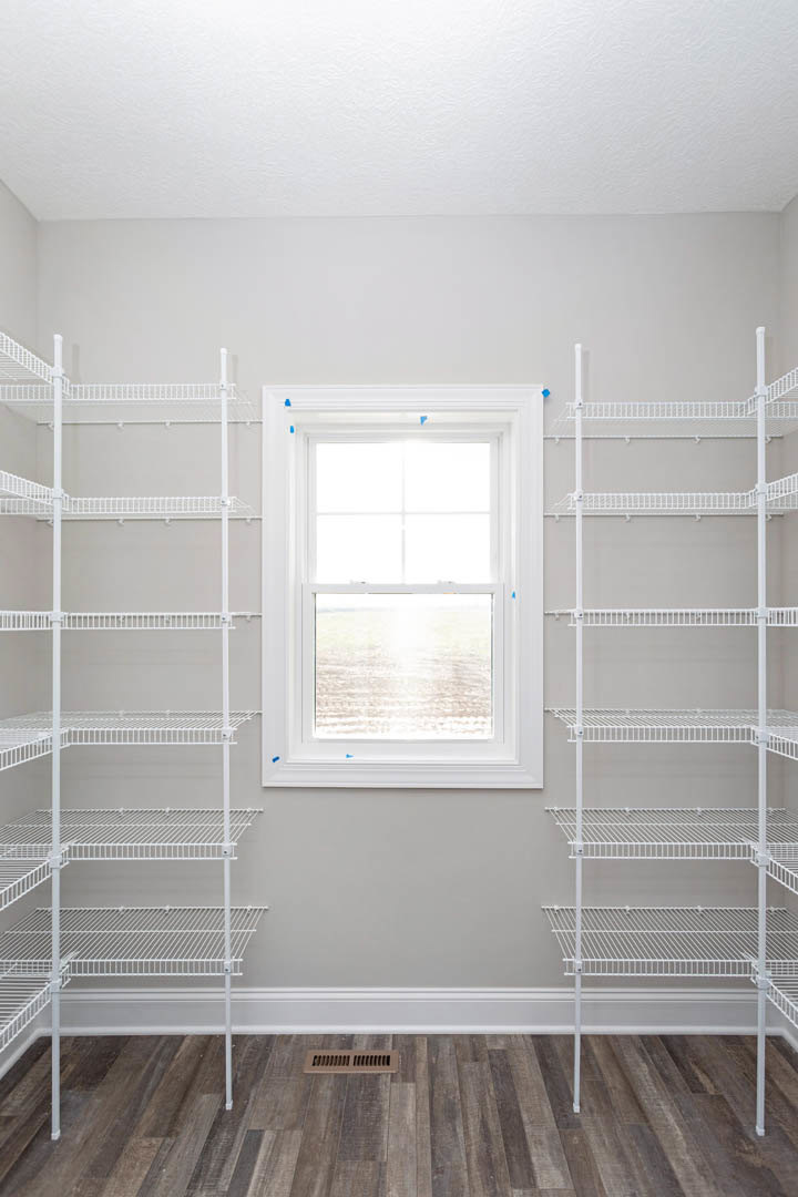 White wire shelving unit along a wall, wood floor with floor vent, large window overlooking grassy field, neutral painted walls