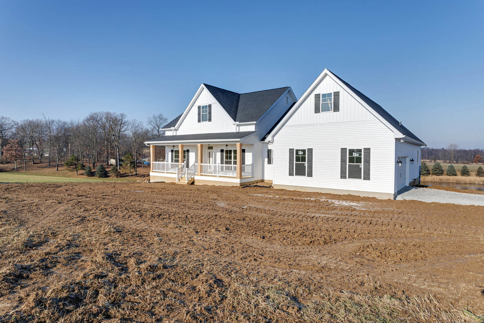 White farmhouse with black roof and white porch railing, set beside a dirt field and bordered by a forested area.
