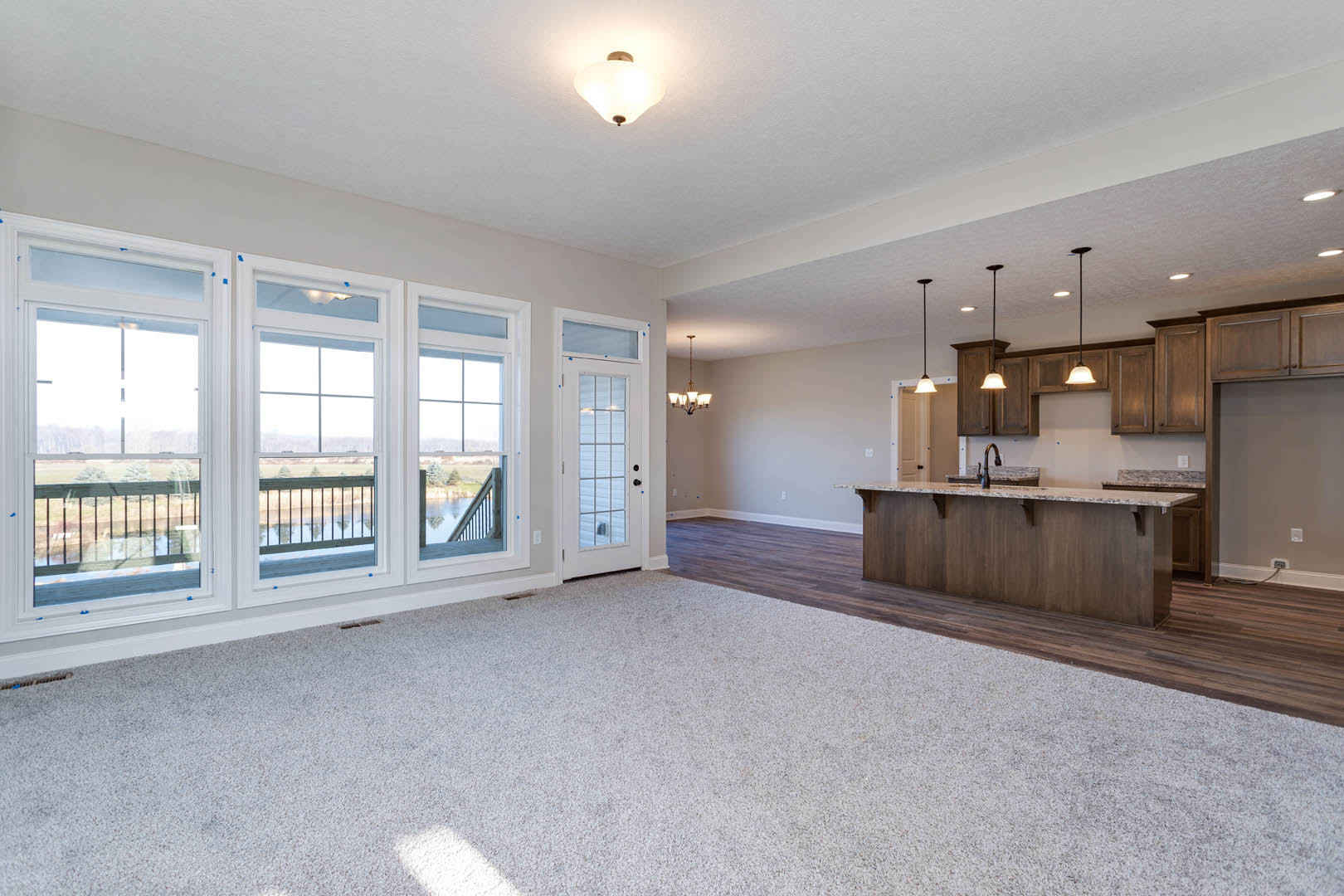 Open living room with carpeted floor, marble kitchen countertop, large windows, and glass door leading to deck with metal railing