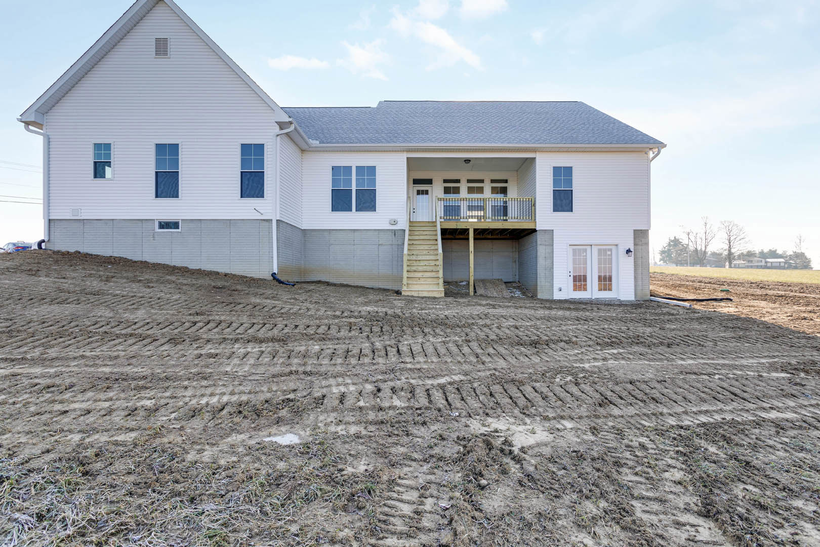 Modern two-story house with light siding, elevated porch, balcony, and square windows, set on a dirt hill with a ramp and staircase leading to the garage.