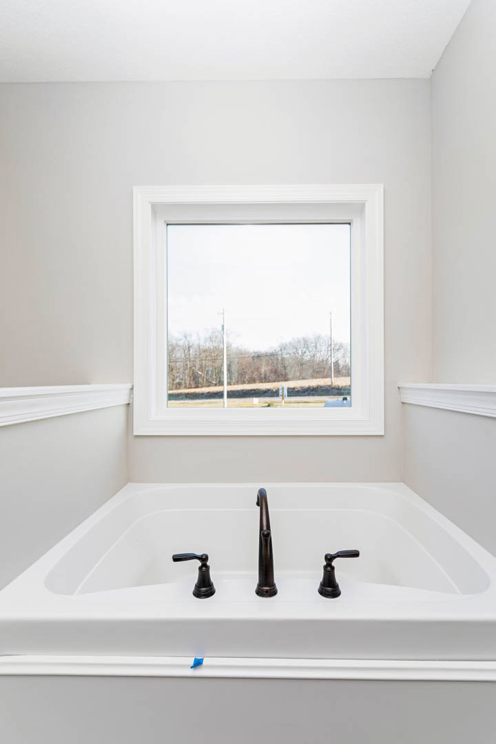 Freestanding white bathtub beneath a large window overlooking a field and trees, chrome faucet mounted on tub edge, neutral bathroom walls and black handle on adjacent white