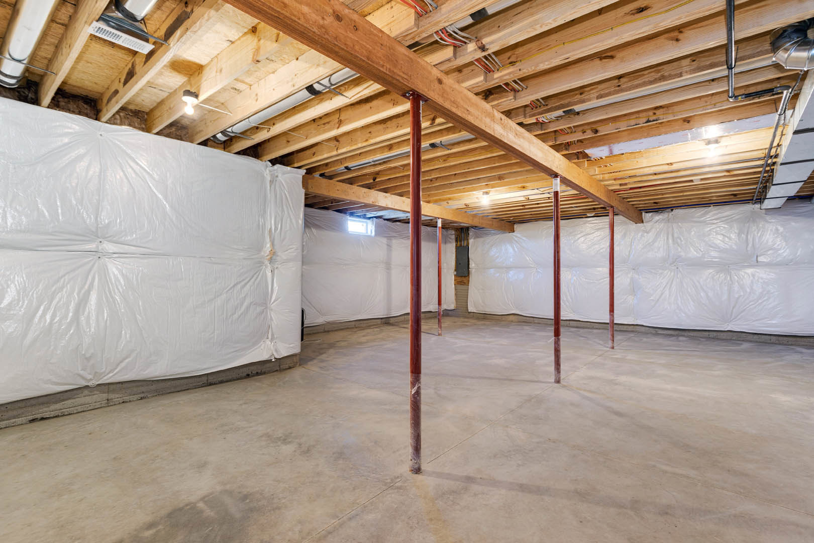 Room under construction with exposed wooden ceiling beam, electrical wires, concrete floor, and walls covered in white plastic sheeting; remote control device visible in