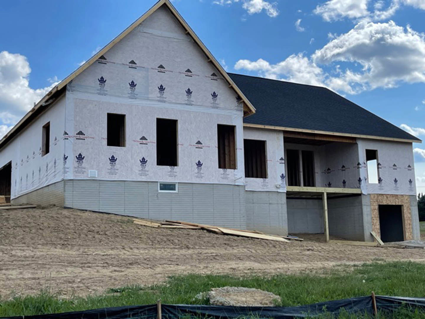 Wood-framed house under construction on a grassy hill, scattered lumber and rocks in foreground, Rockingham Meeting House visible in background, blue sky with clouds overhead