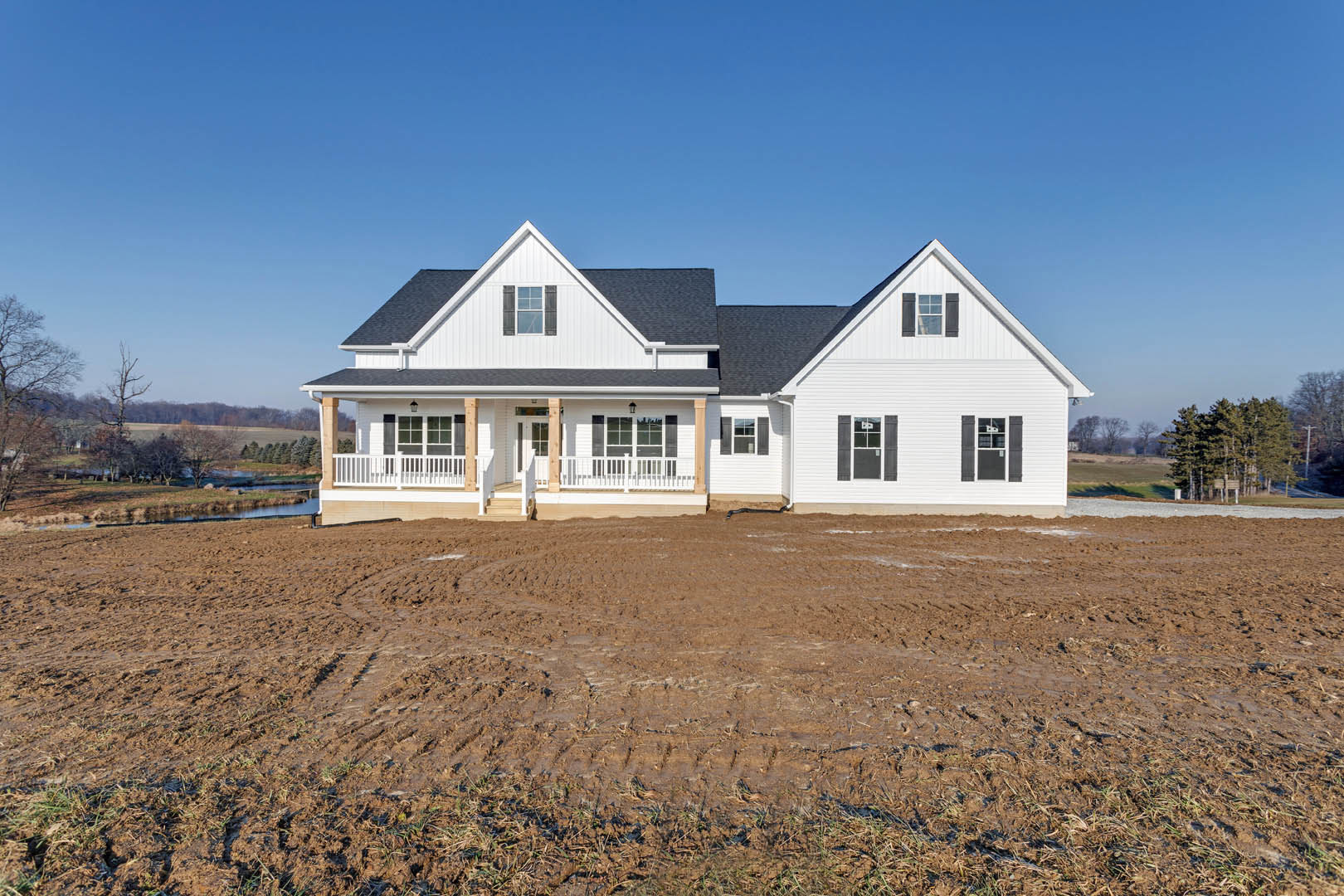 White farmhouse with covered porch, muddy yard featuring tire tracks, Southfork Ranch visible in background, blue sky, scattered trees along property edge.