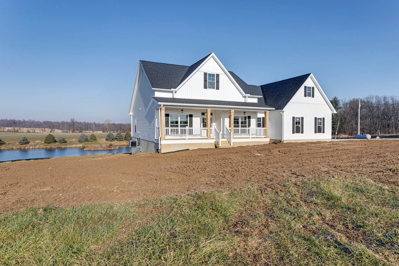 White farmhouse with black roof and white-framed windows, surrounded by grass and dirt field, overlooking a pond with trees and Southfork Ranch visible in the distant background.