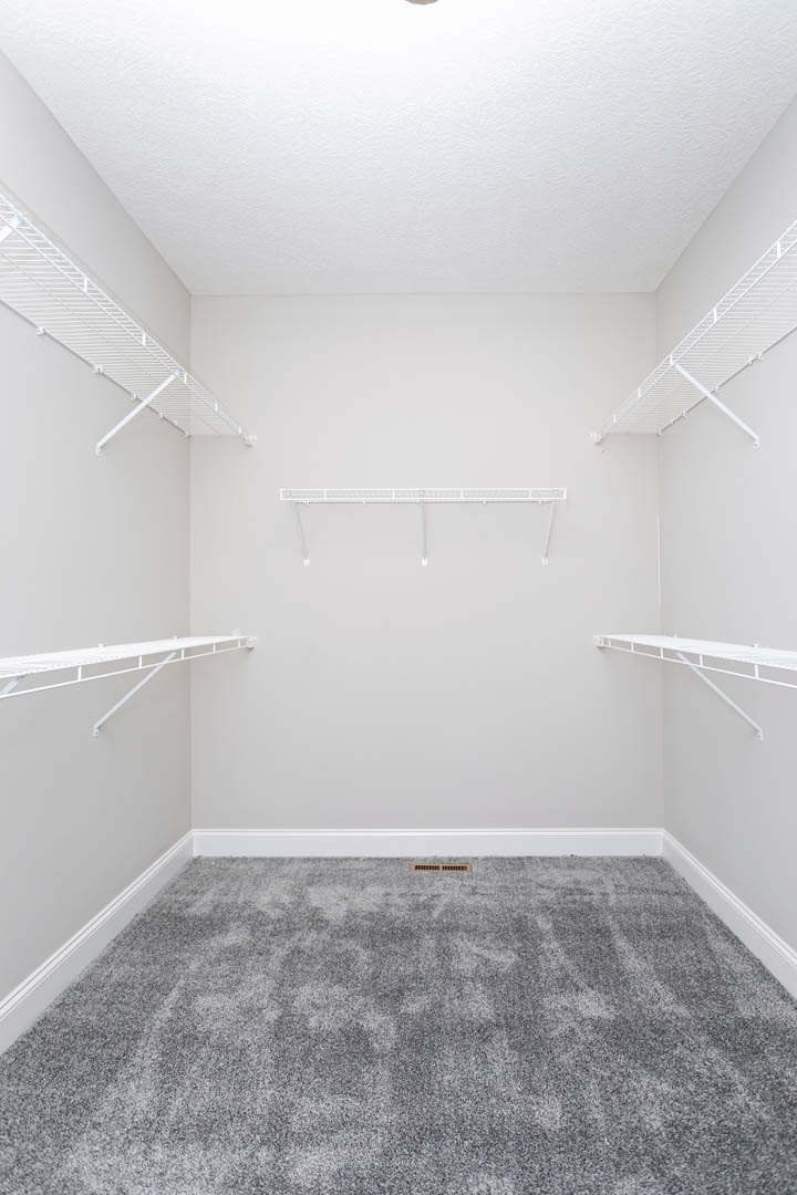 White built-in shelves against a white plaster wall, grey carpeted floor with a vent, recessed ceiling light, minimalist interior design.