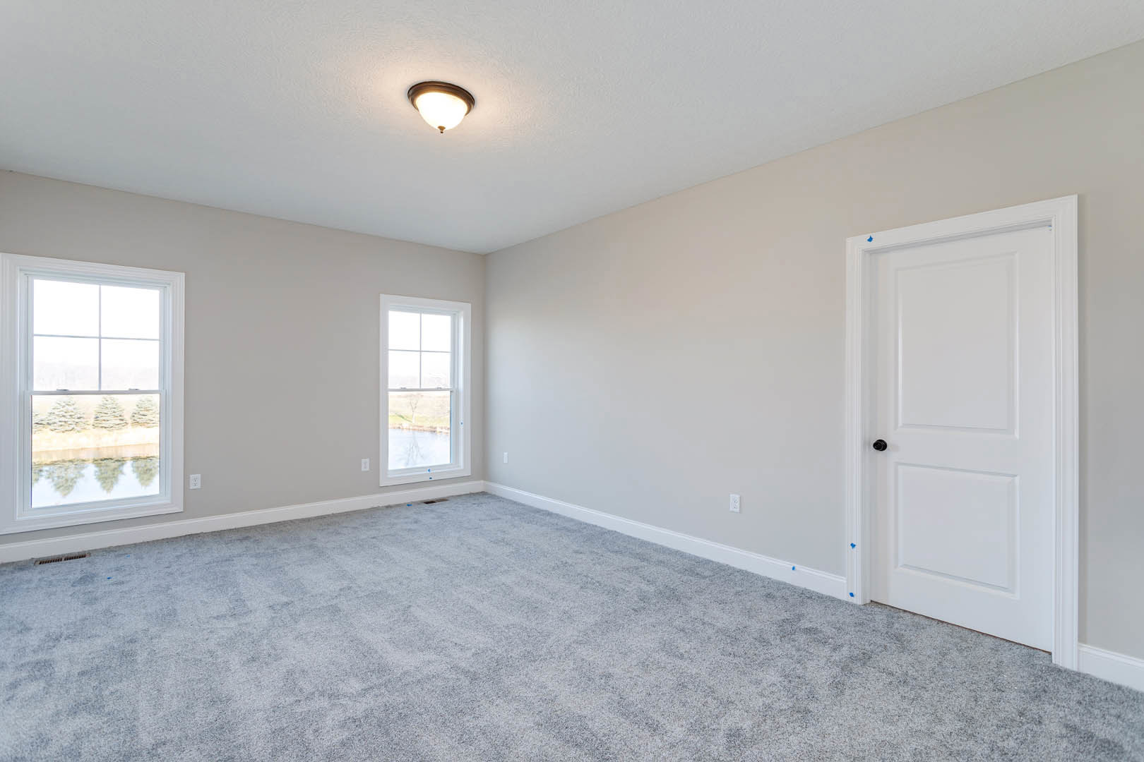 Carpeted room with white walls, white door with black knob, ceiling light fixture, large windows offering views of river, trees, and lake