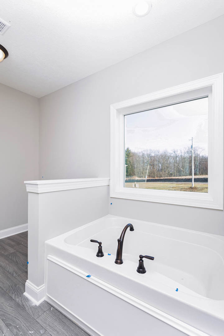 Freestanding white bathtub with chrome faucet beneath large window overlooking grassy field and power lines, light-colored bathroom walls and floor visible