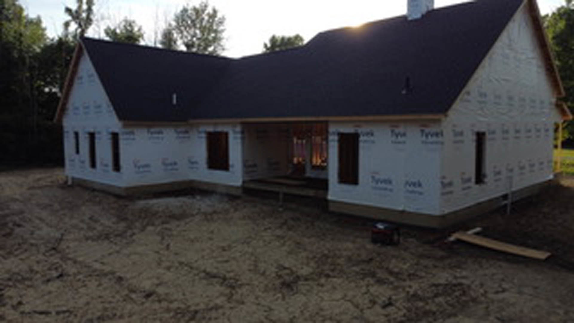 Modern house under construction with black roof, exposed grey foundation, open doorway, dirt ground, and surrounding trees