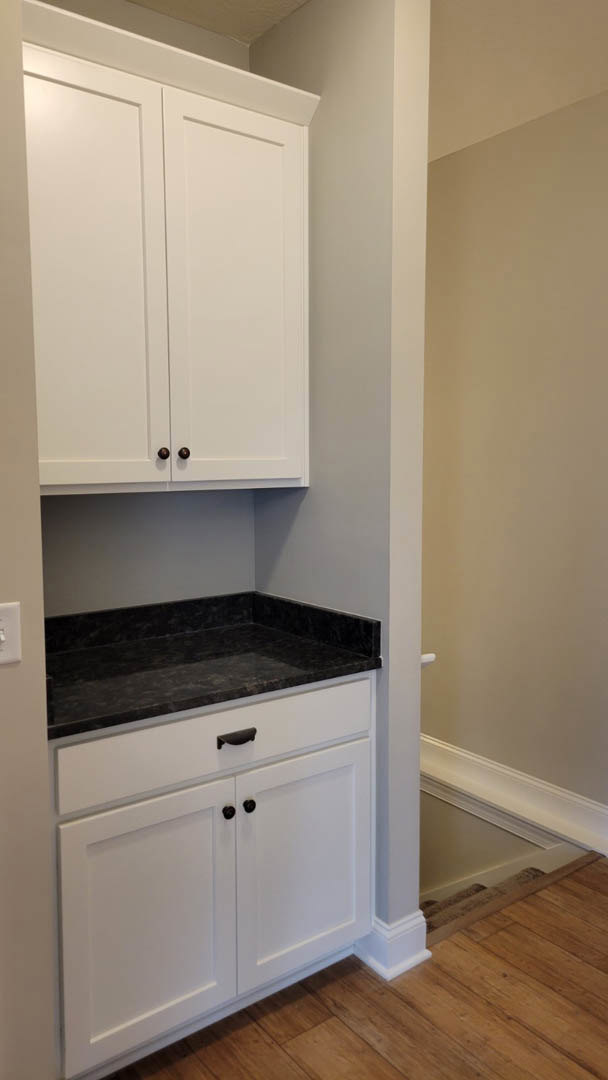 White kitchen cabinets with black countertops, wood flooring, black cabinet handles, and white walls