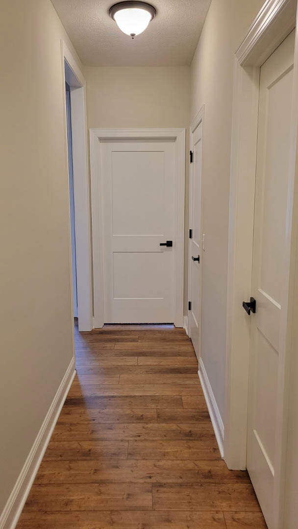 Hallway with warm wood flooring, white paneled doors featuring black handles, white baseboards, and a modern ceiling light fixture