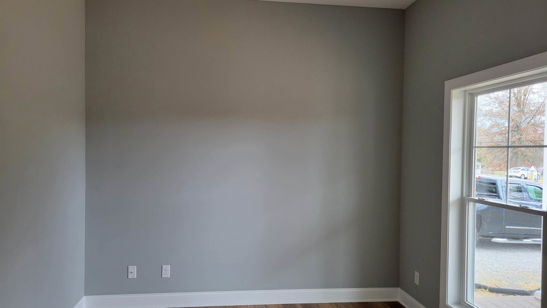Gray-walled room with white ceiling, large window showing parked car and tree outside, minimal interior finishes