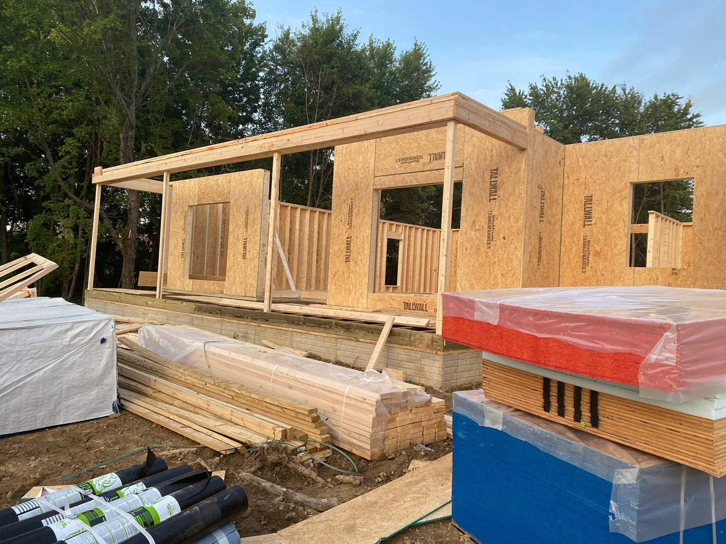 Framed house under construction with exposed wooden beams, stacks of lumber, black and white pipes, white tarp covering part of the structure, and red foam mattress wrapped in