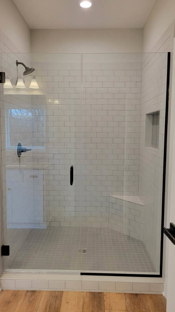 Walk-in shower with clear glass door, chrome shower head mounted on white tiled wall, linear drain on light wood floor, recessed ceiling light fixture