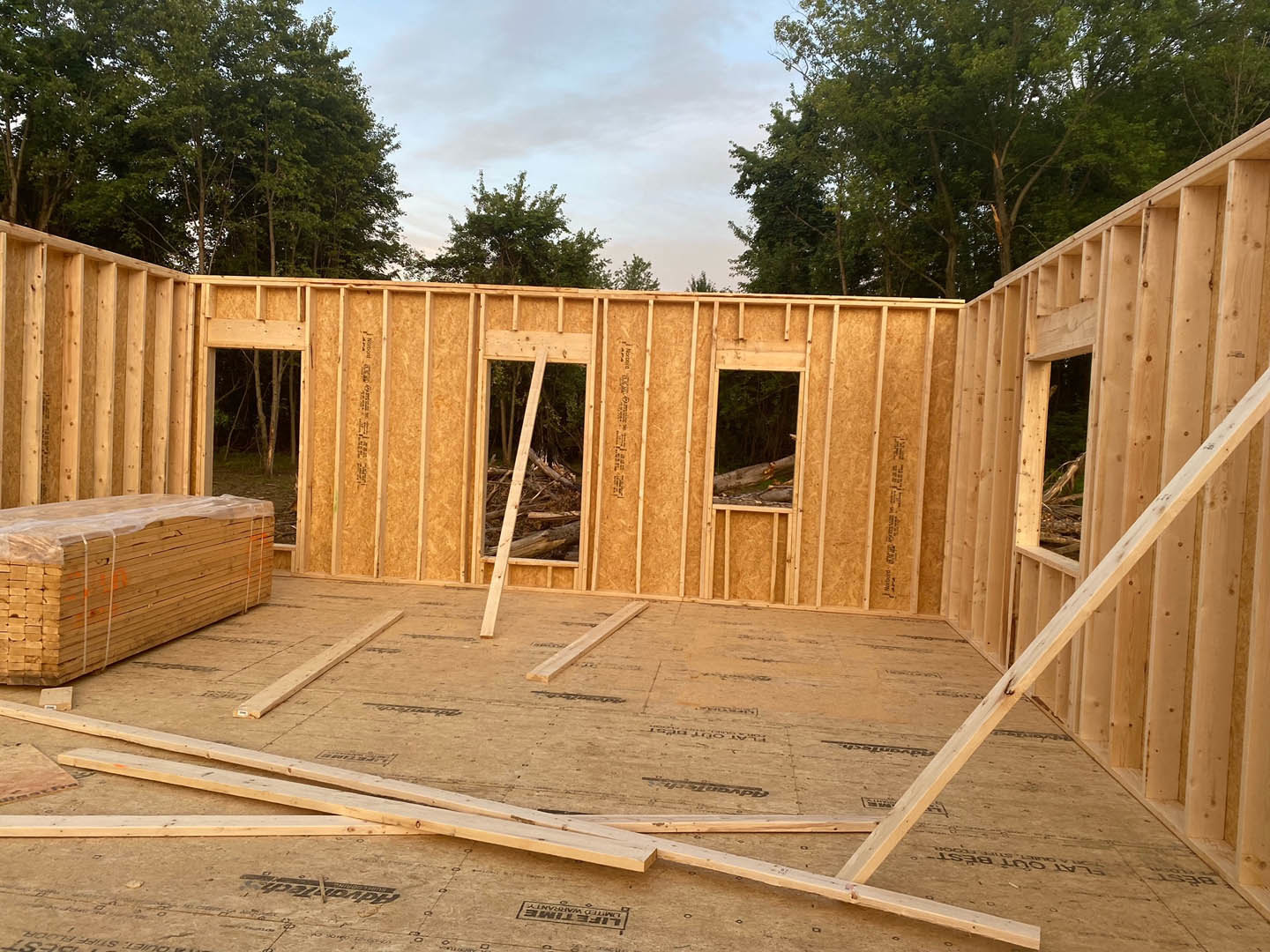 Wood-framed house under construction with stacked lumber wrapped in plastic, wooden planks on the floor, broken window frame, and wooden railing; blue sky and trees in the