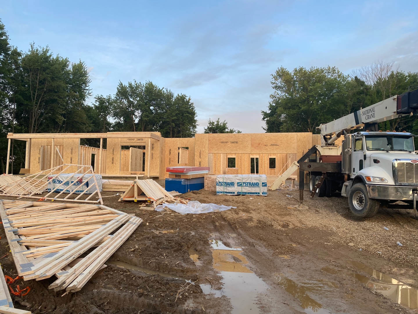 Construction site with a truck parked in front of a partially built custom home, wood planks stacked nearby, crane mounted on truck, white sign with blue text, trees and blue sky