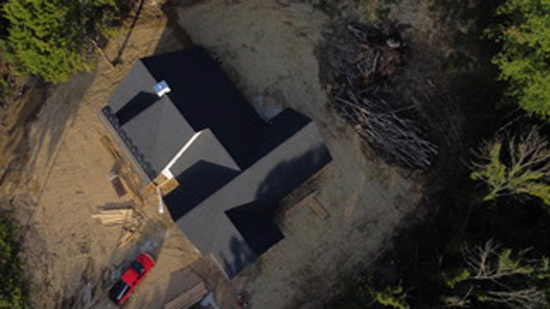 Two-story custom home with black metal roof, white siding, large windows, and a red car parked in front; mature tree with green leaves in foreground.