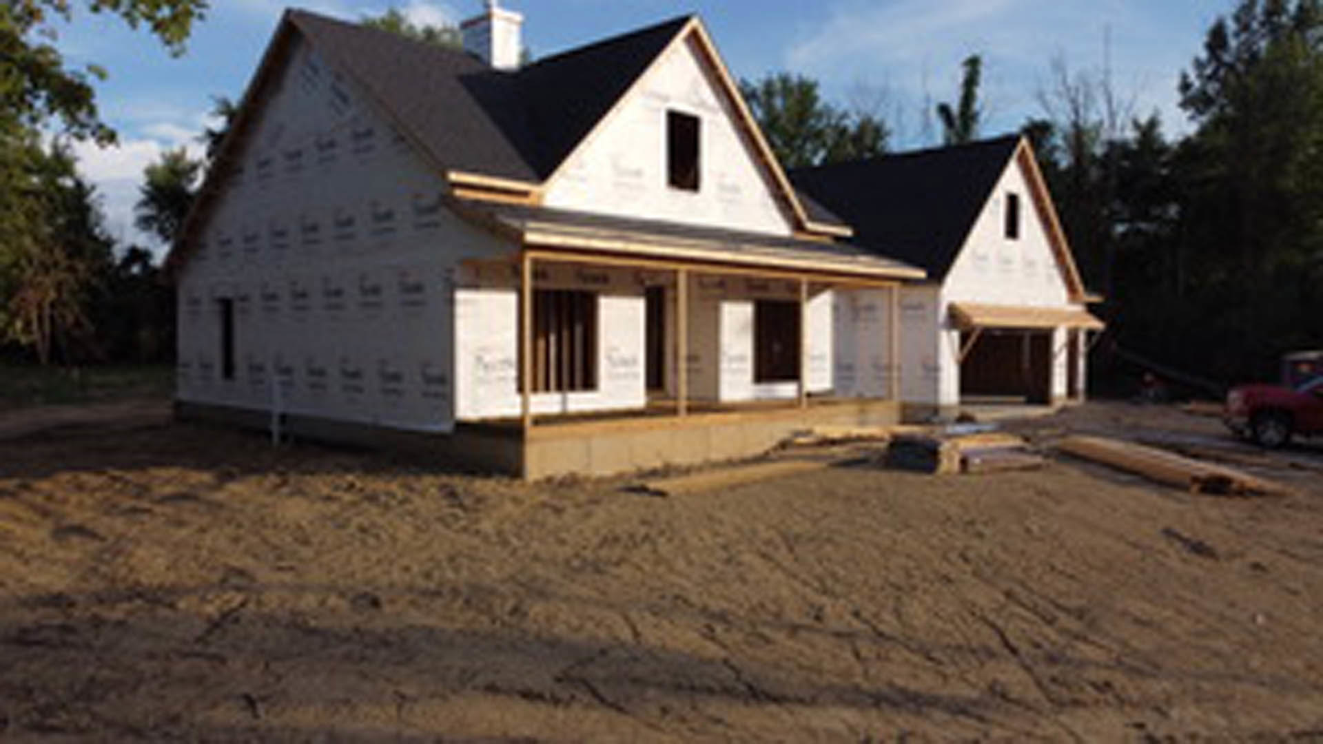 Wood-framed house under construction with exposed beams and partially finished porch, surrounded by dirt lot and scattered construction materials, trees and blue sky in background