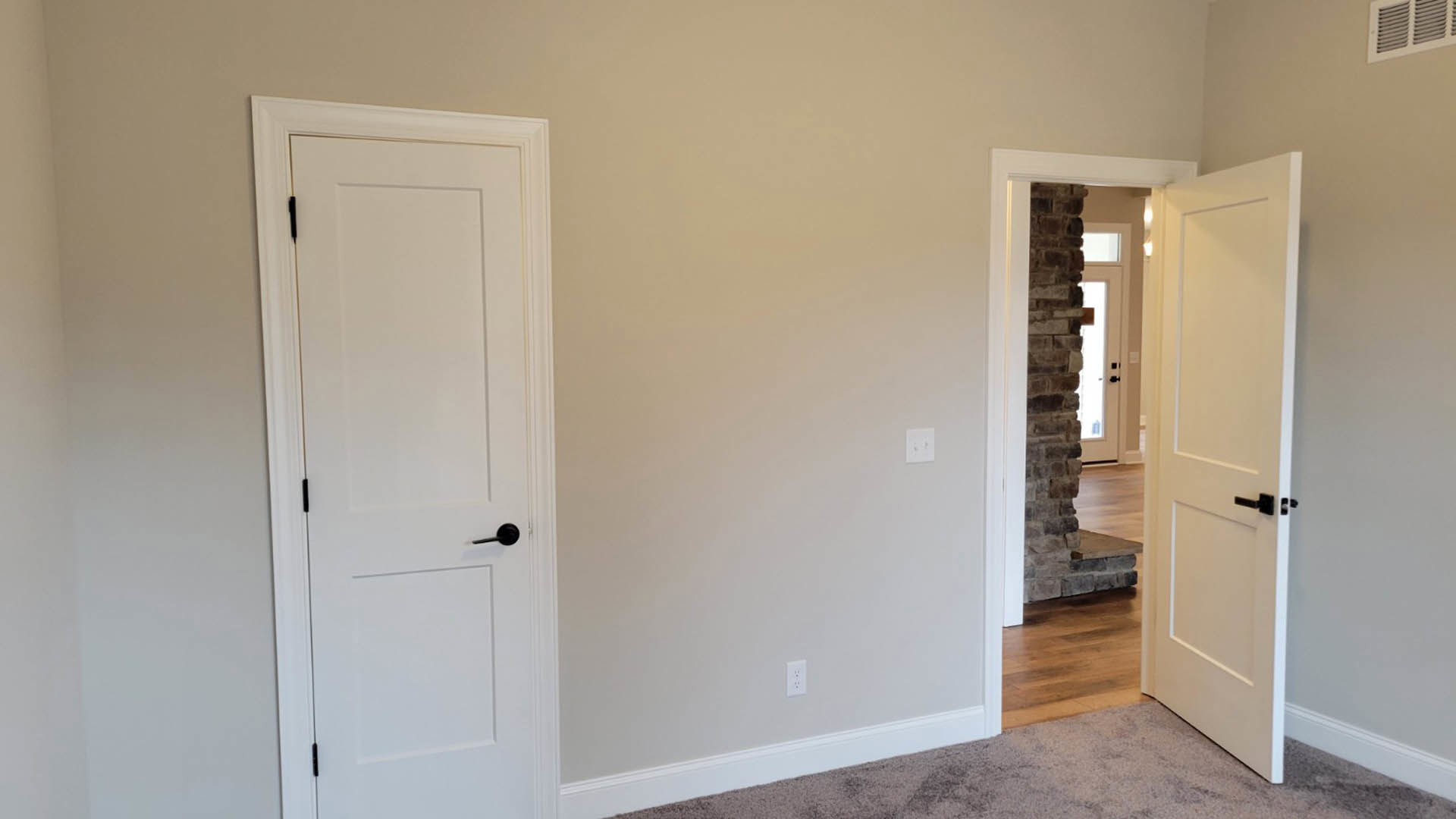 White paneled door with matte black handle, light switch on adjacent white wall, beige carpet flooring, rectangular air vent near baseboard