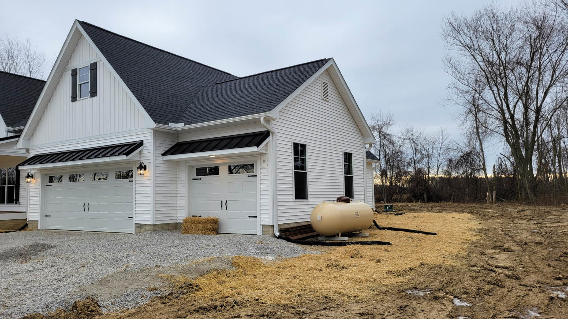 White house with black roof and white-framed window, large tan tank elevated on metal stand in front, leafless trees in background, overcast sky.