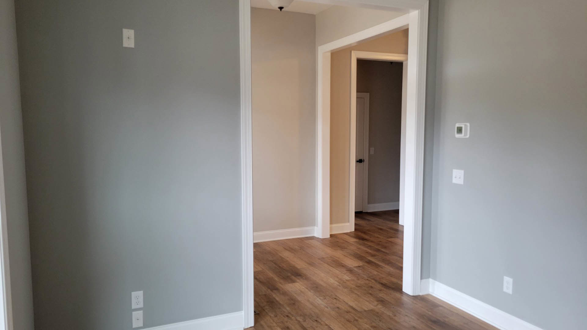 Hallway with wood flooring, white walls, ceiling light fixture, white rectangular clock with black star, wall-mounted light switch and outlet with silver knobs