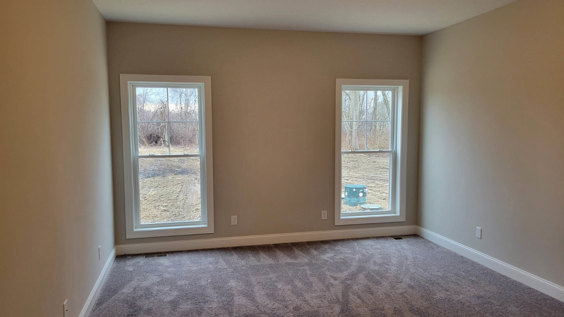 Bright room with two large windows, light plaster walls, neutral carpet flooring, blue cylindrical stool near a window, partial view of tree and sand outside