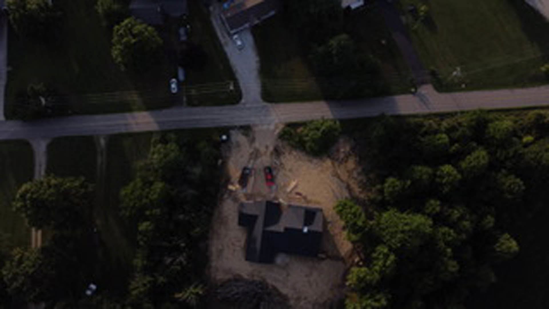 Modern house with black roof surrounded by bare dirt, bordered by mature trees, with a dirt road and neighborhood street visible in the background.