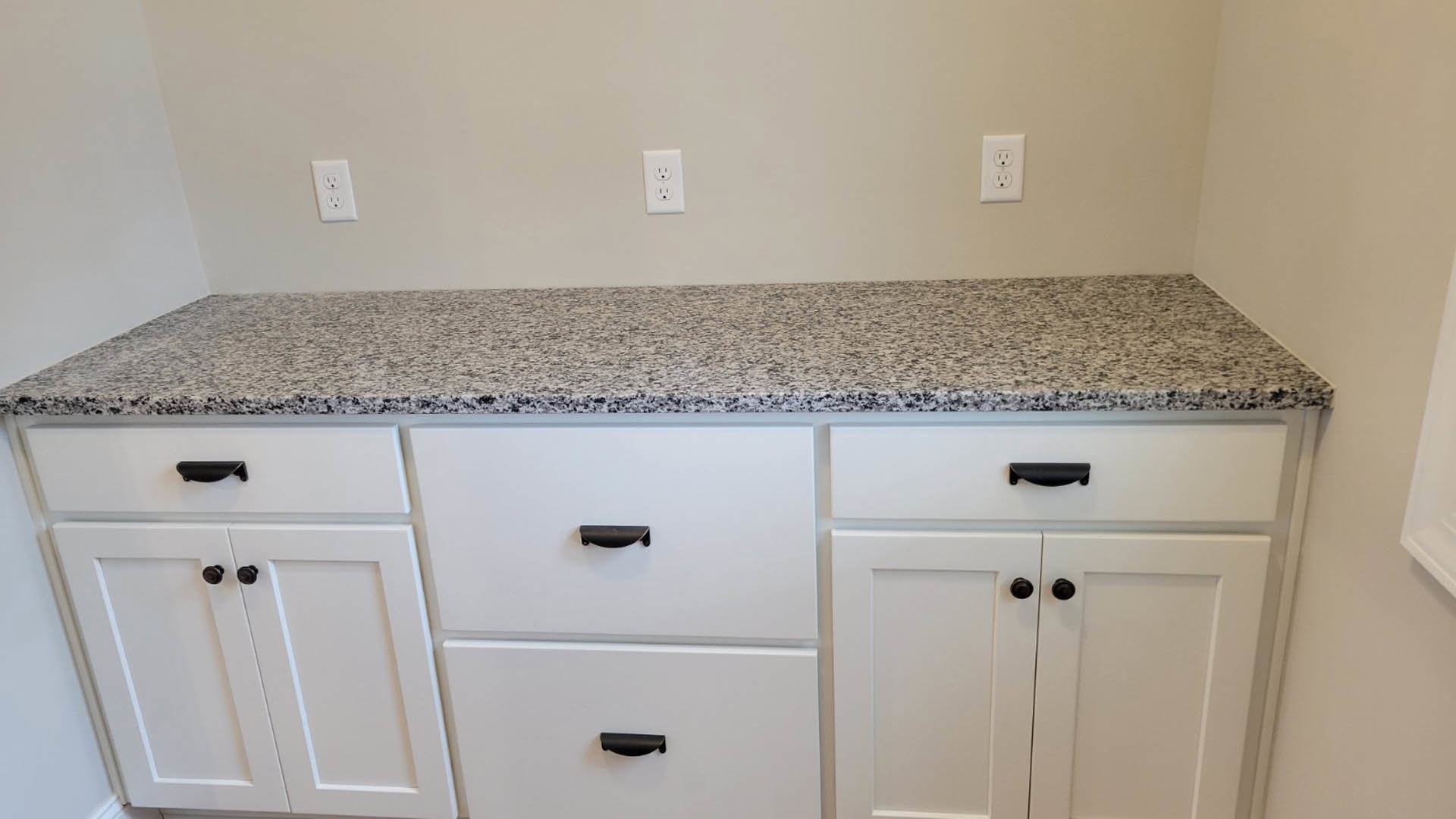 White kitchen cabinets with black handles beneath a granite countertop, featuring drawers and a visible electrical outlet.