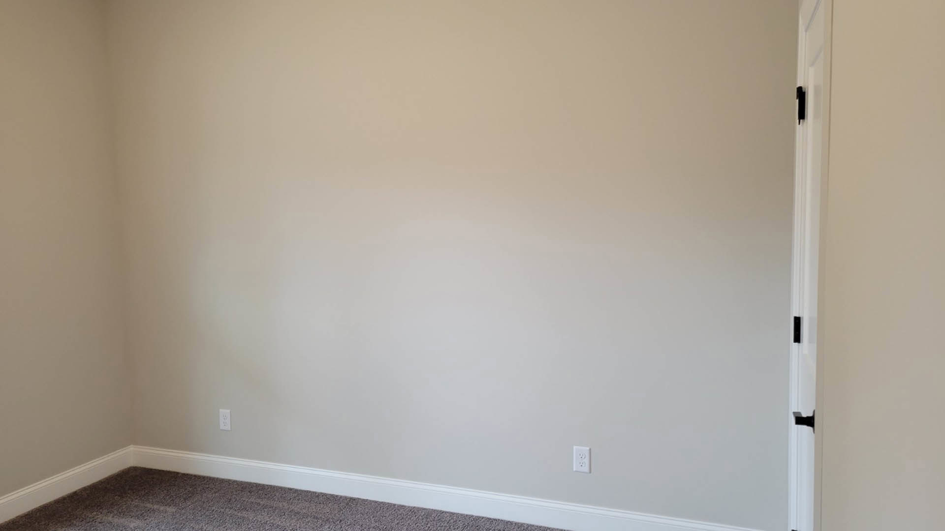 White plaster wall with white electrical outlet, beige carpet flooring, close-up of silver door handle on brown door, white door frame.
