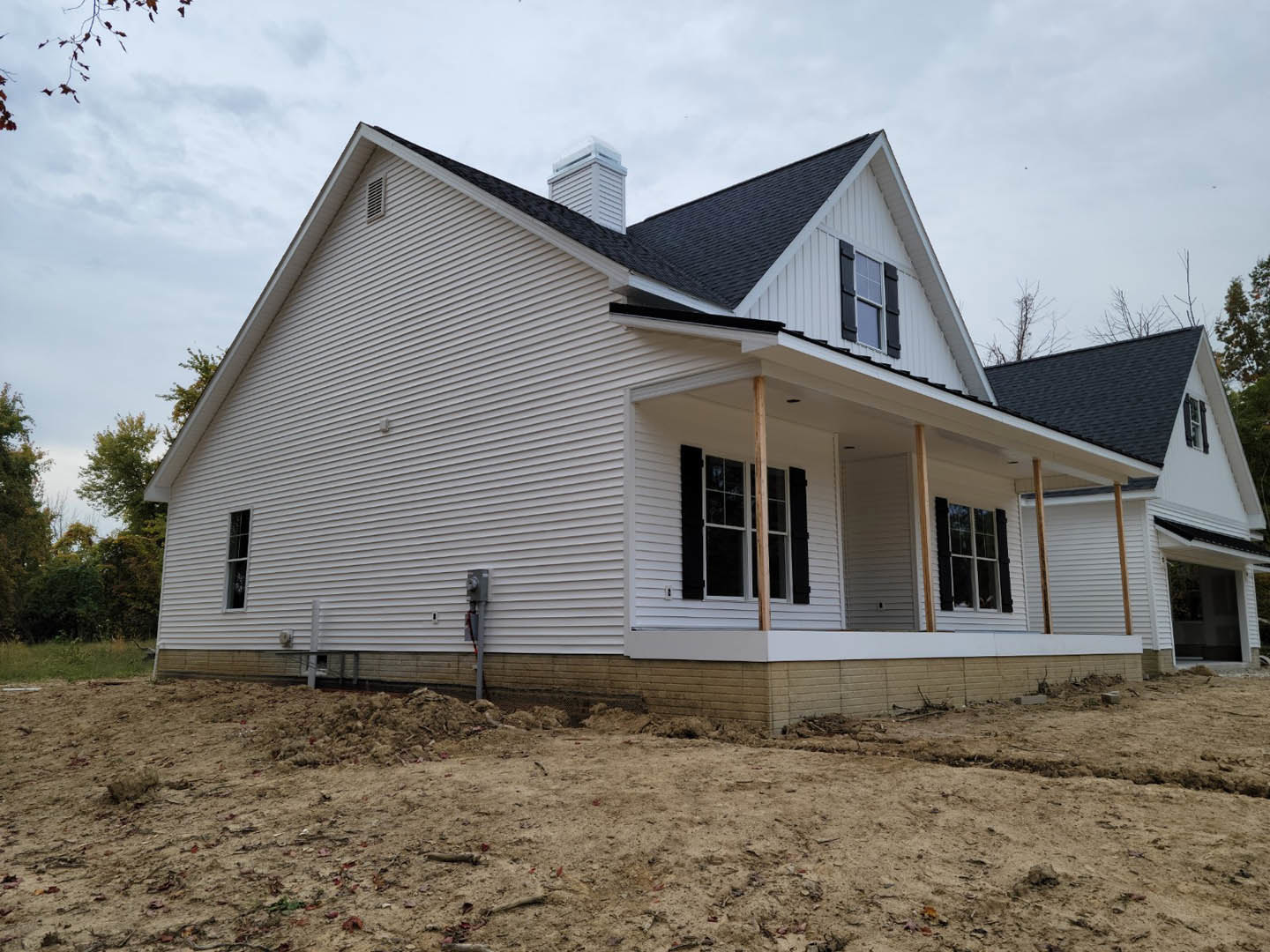 White house with black shutters under construction, dirt ground with debris pile, surrounded by trees, visible porch and windows, gray roof, cloudy sky overhead
