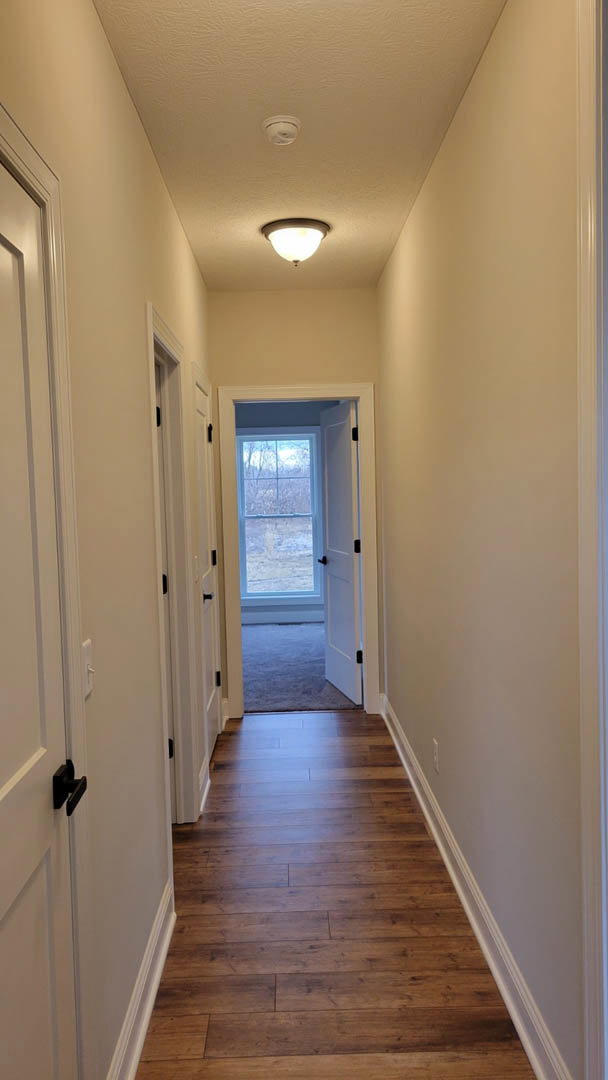 Hallway with white plaster walls, wood laminate flooring, ceiling-mounted light fixture, open white door with black handle, window featuring a blue frame
