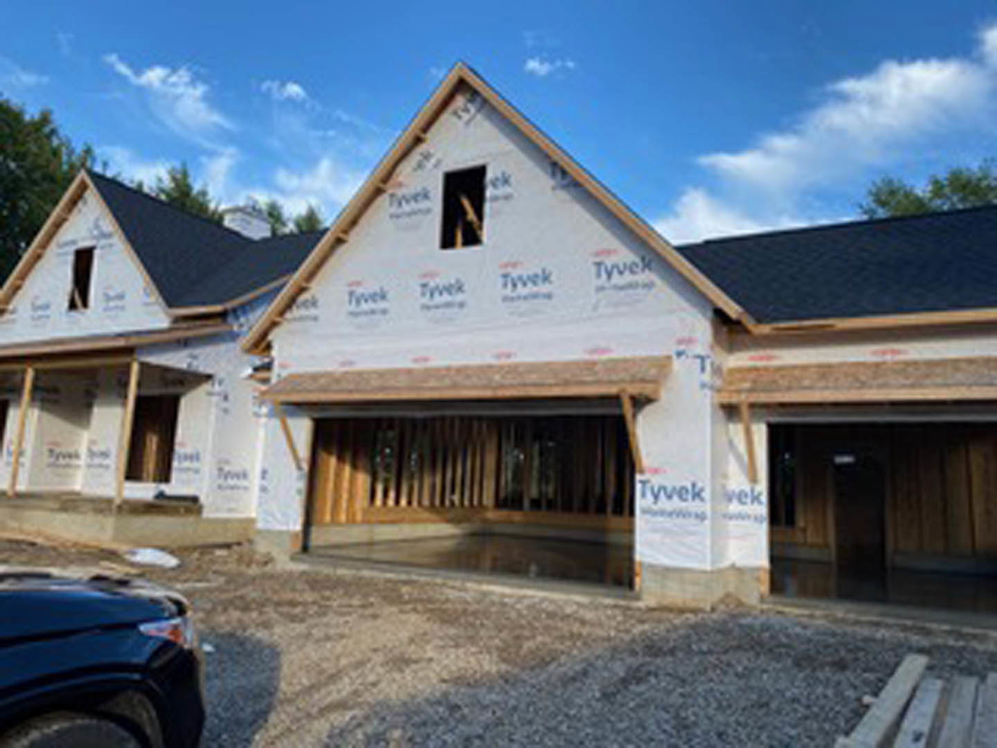 Two-story house under construction with exposed wooden framing, attached garage, black car parked in driveway, cloudy sky overhead