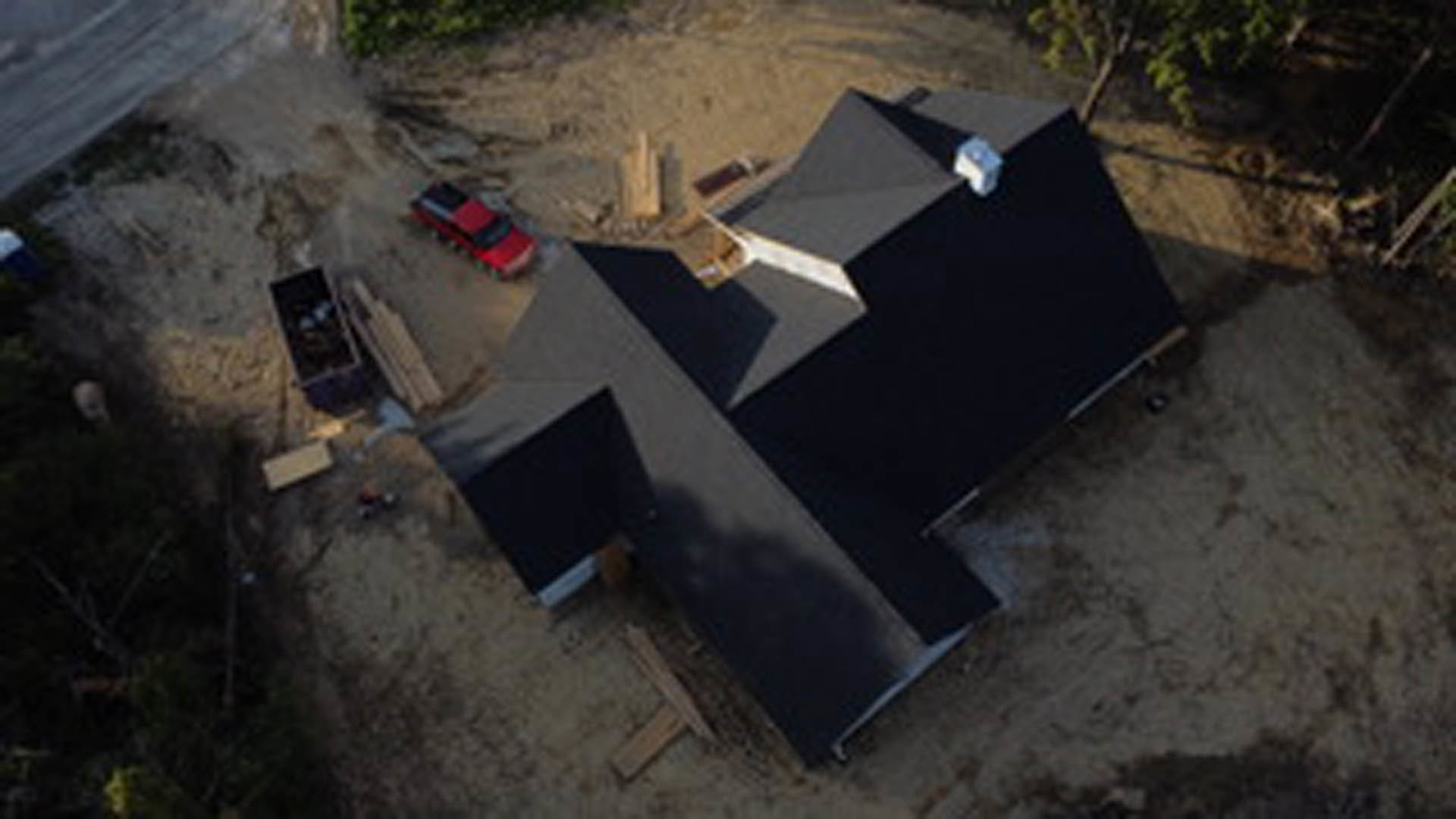 Modern two-story home with black shingle roof, light-colored siding, large windows, and driveway with parked cars; construction materials visible in front yard.