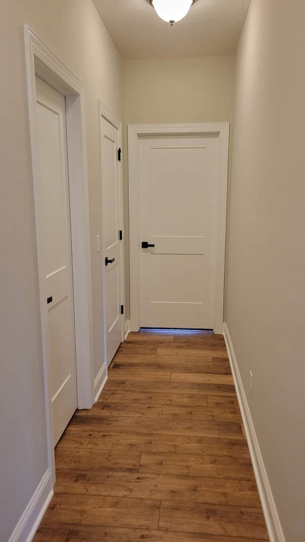 Hallway with white paneled doors featuring black handles, wood plank flooring, white walls with white trim, illuminated ceiling light fixture