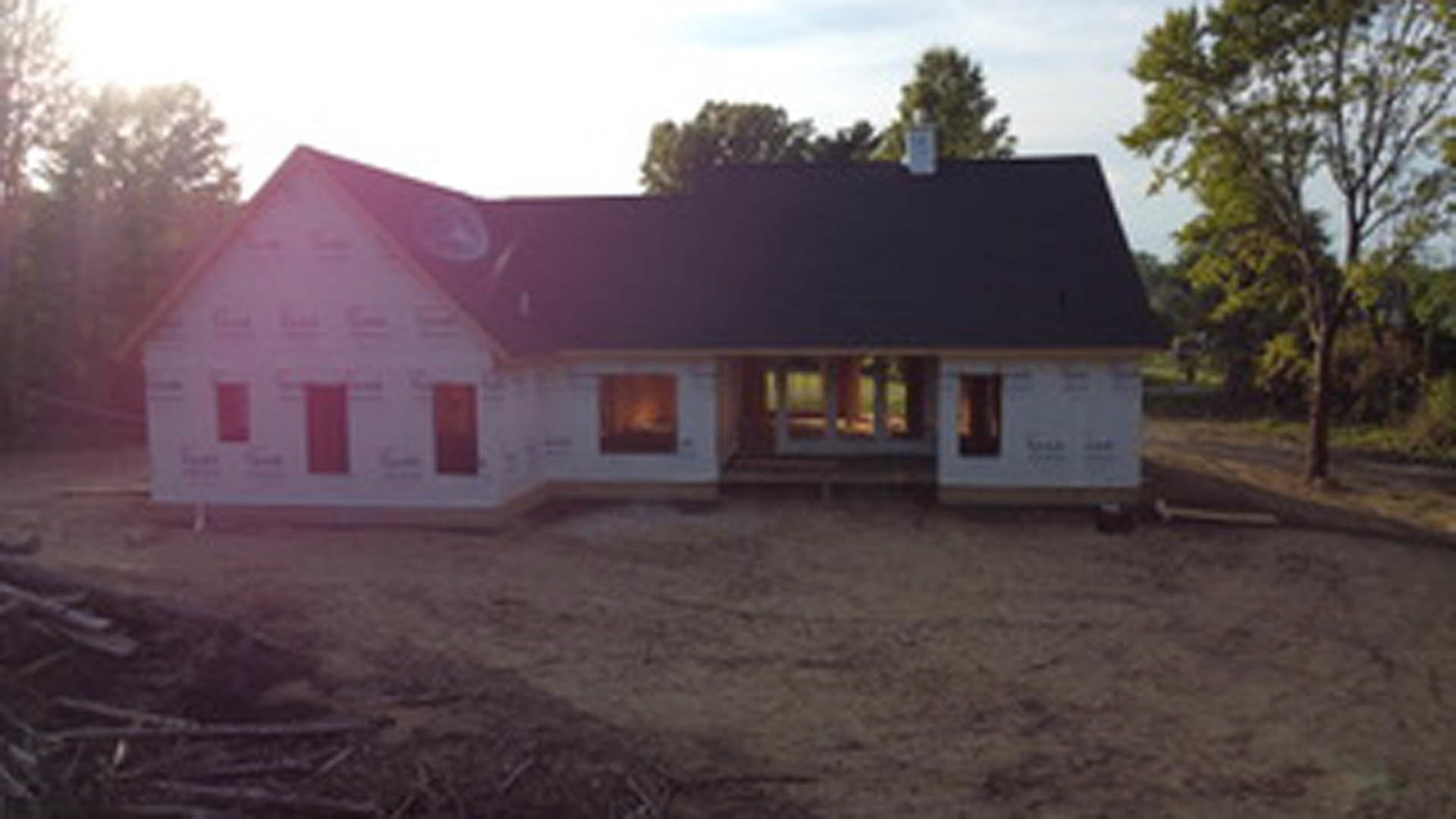 Partially built house with black roof, white exterior walls, exposed dirt ground, surrounded by tall trees in background