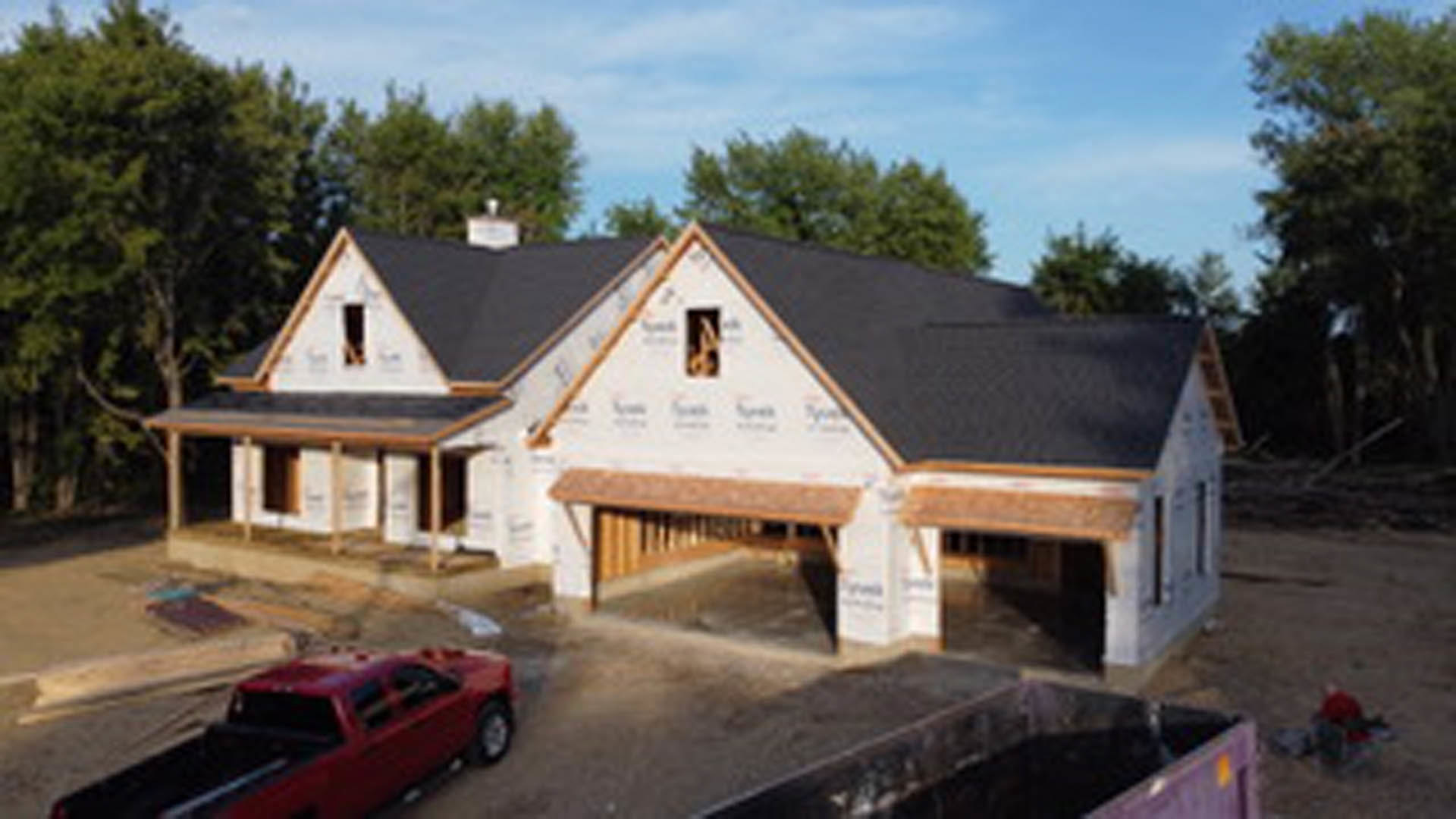 Partially built house with exposed framing, unfinished exterior walls, and a red car parked in the driveway; surrounding area includes trees and open sky.