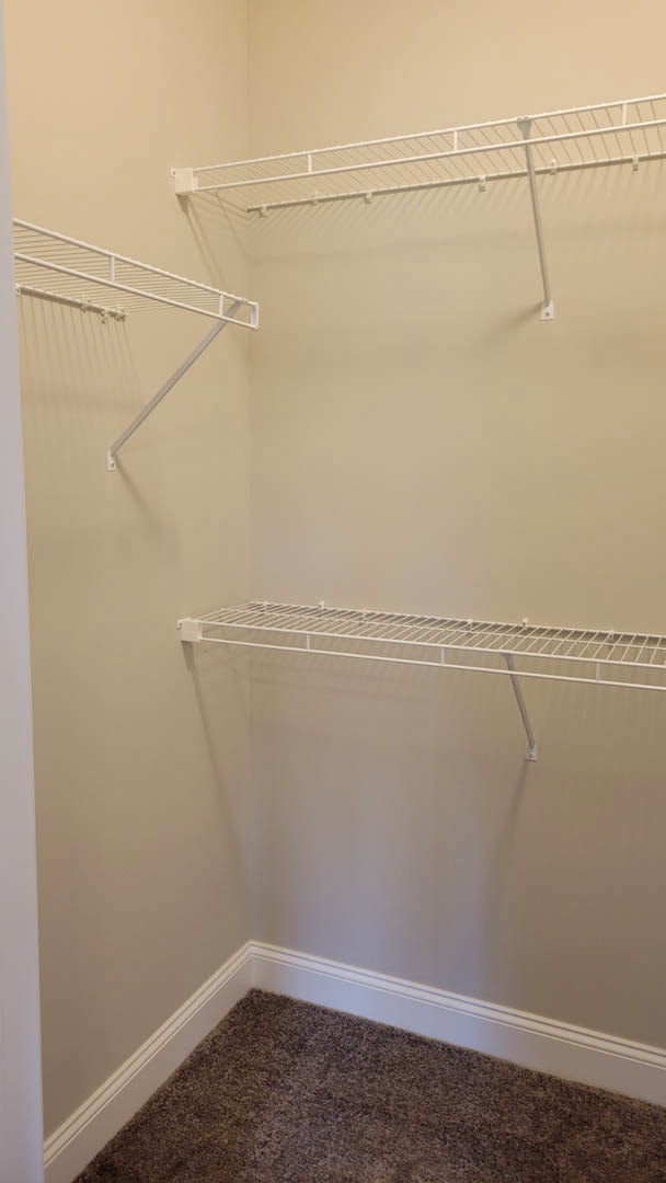 White built-in shelves with metal bars in a closet, white plaster walls, carpeted floor, and a white railing along the wall.