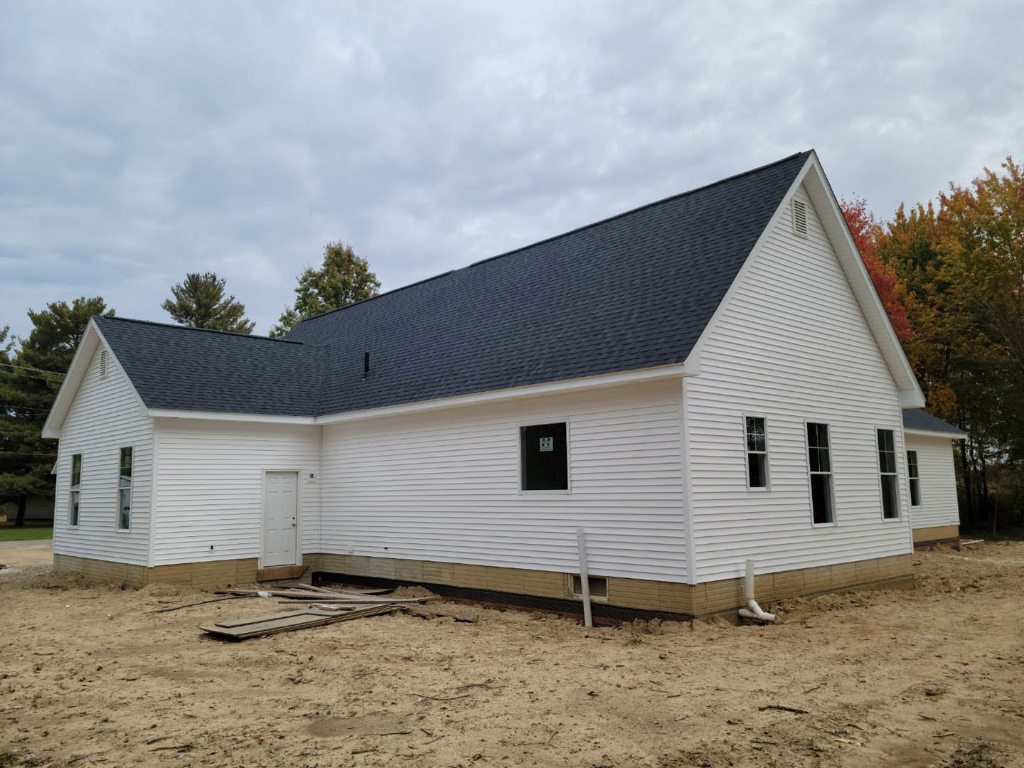 White house with black roof and black door, white siding, dirt yard with scattered debris, trees and cloudy sky in background
